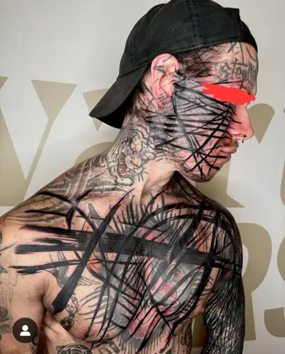 A heavily tattooed man wearing a black cap is shown in a side profile. His face, neck, and upper body are covered in intricate black linework and abstract tattoo patterns, layered over older tattoos. His eyes are redacted with a red mark. The background features a white wall with large, partially visible gold letters.